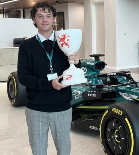 Male student stands next to Formula One race car holding racing cup trophy.
