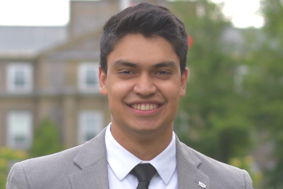 Drabble dressed formally, smiling and holding a black folder on campus. 