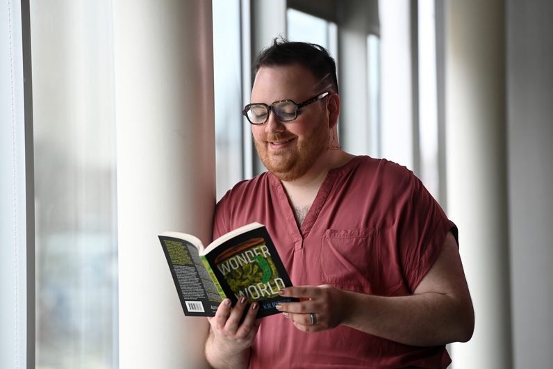 K.R. Byggdin leans against a white post smiling and looking at the open book in their hands, which is black and green and titled "Wonder World."