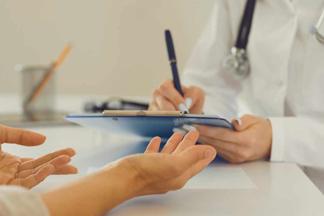 The hands are shown of one person open and gesturing while another in a lab coat and stethoscope on their shoulder writes on a clipboard. 