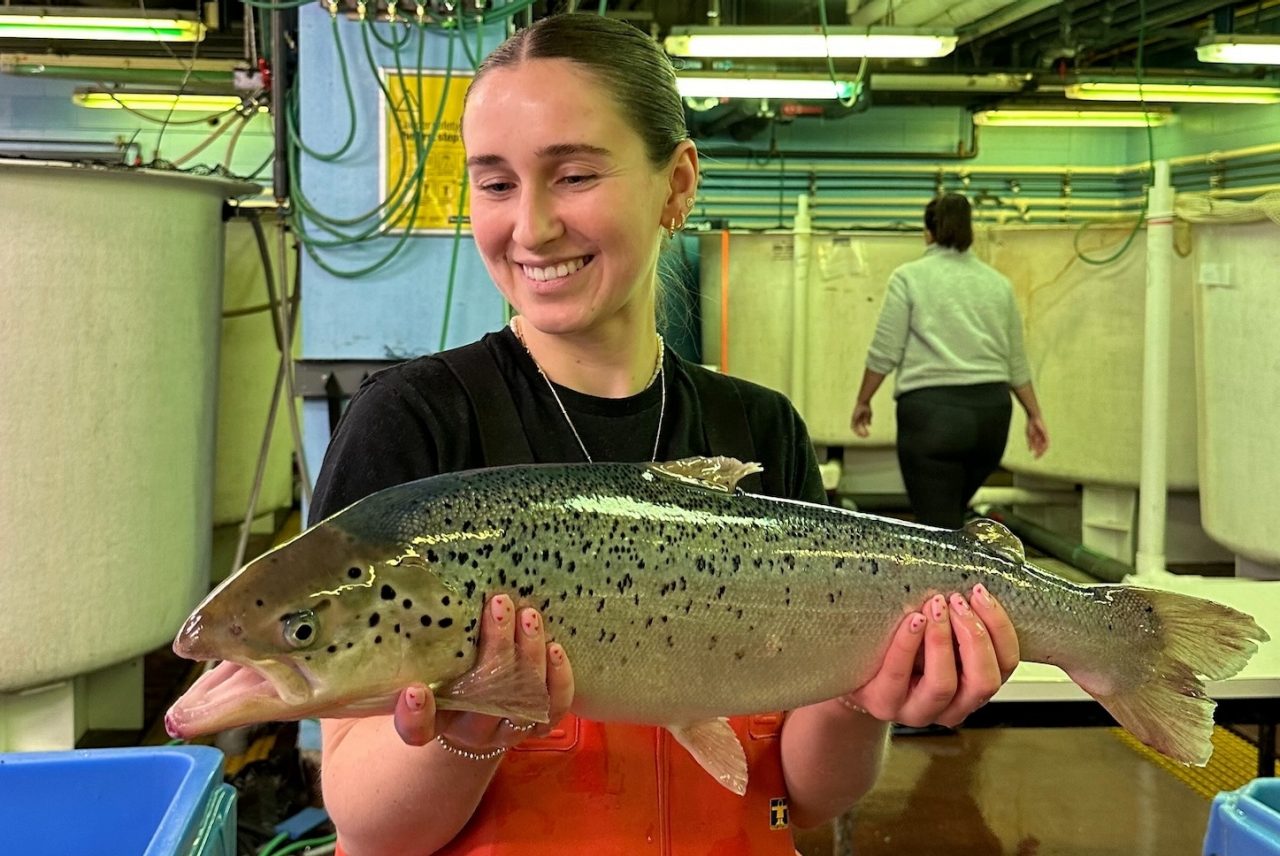 Sernowsky wearing orange hip wader overalls and smiling while holding a medium-sized fish inside the Aquatron lab.