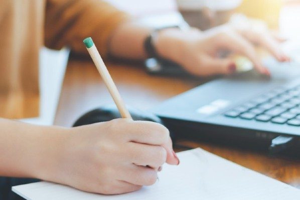 A close-up of a person holding a pencil to paper in one hand with the other hand on a laptop. 