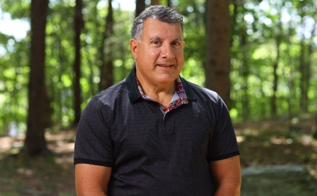 Robert Zed stands outside in a park with trees in the background.