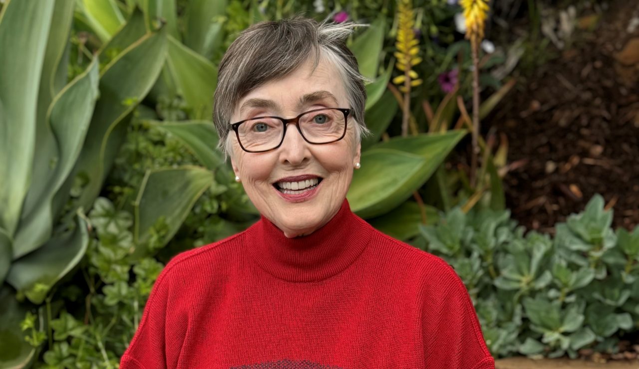 A woman with short grey hair and glasses sits outside in a garden