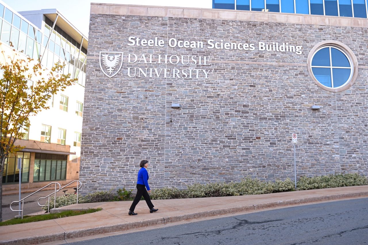 A woman wearing a blue jacket walks in front of the stone Steele Ocean Sciences Building at Dalhousie University. 