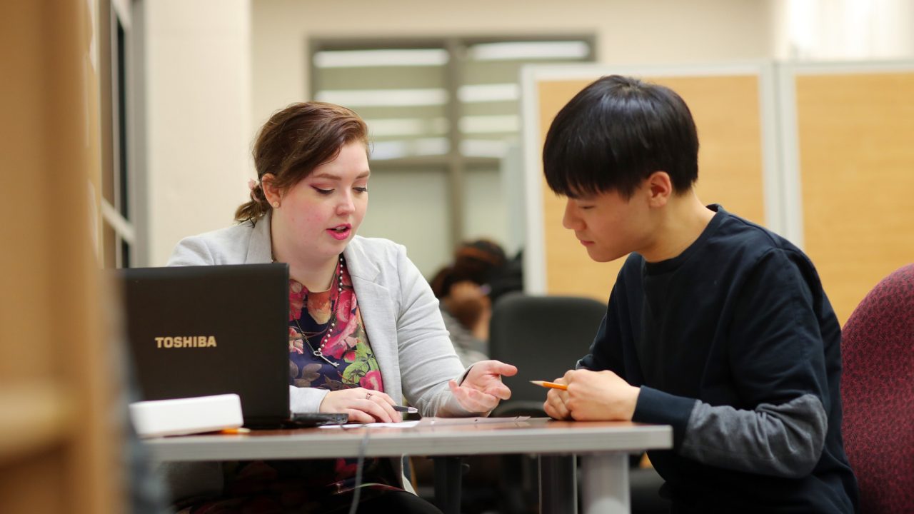 A female-presenting person assists a male-presenting person while seated at a table.