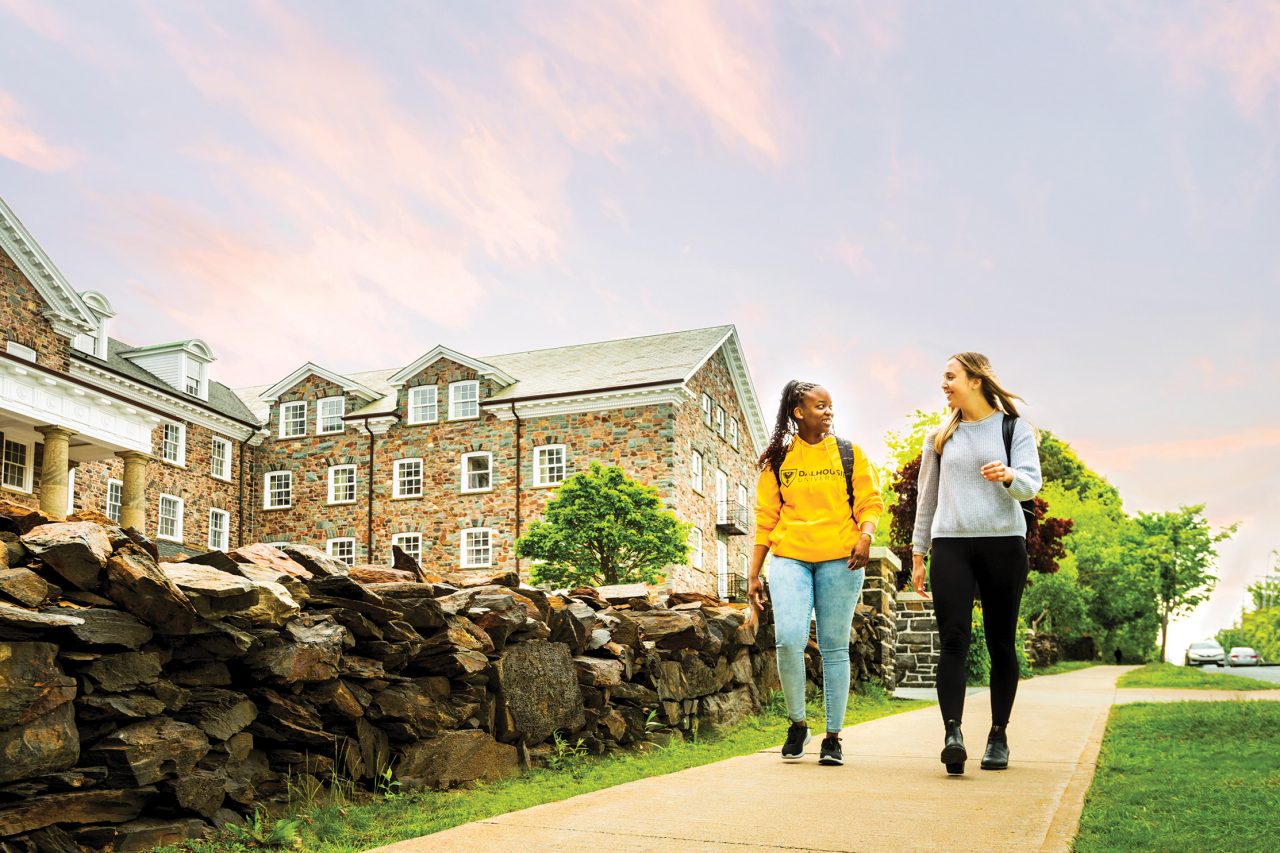 Two female students walk on the sidewalk in front of old stone buildings at sunset. The sky is blue with pink clouds.