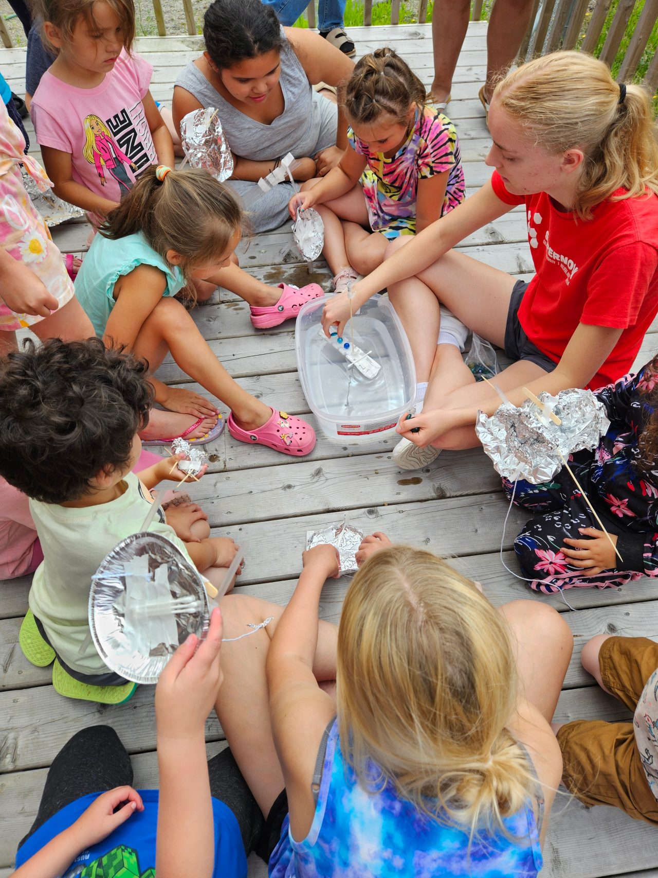 1. A group of children sitting together on a wooden deck, enjoying their time outdoors in a sunny environment.
