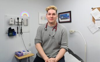 Tanas Silliboy sits in a patient exam room with a stethoscope draped around their shoulders.