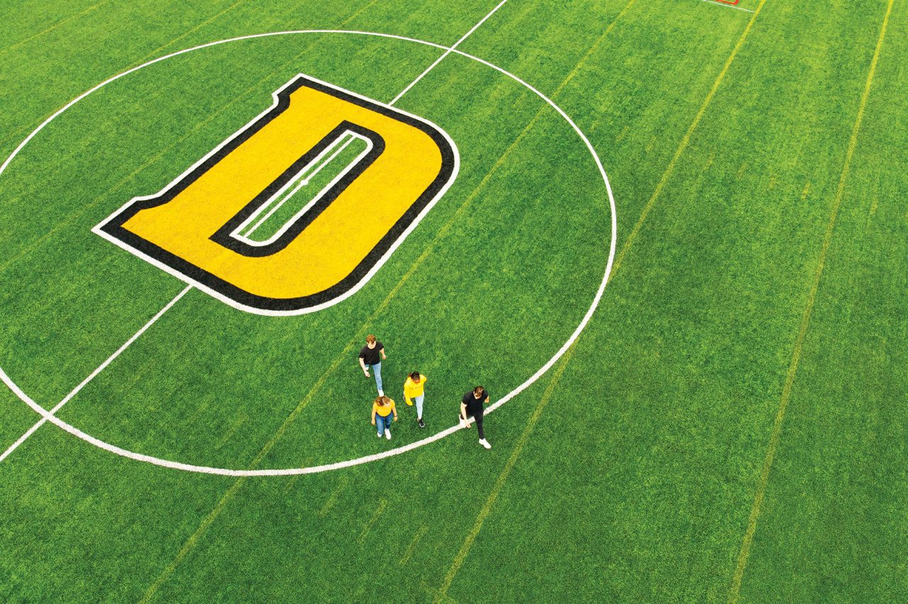 Aerial view of four students wearing black and yellow crossing green sports field painted with a large D surrounded by a white circle.