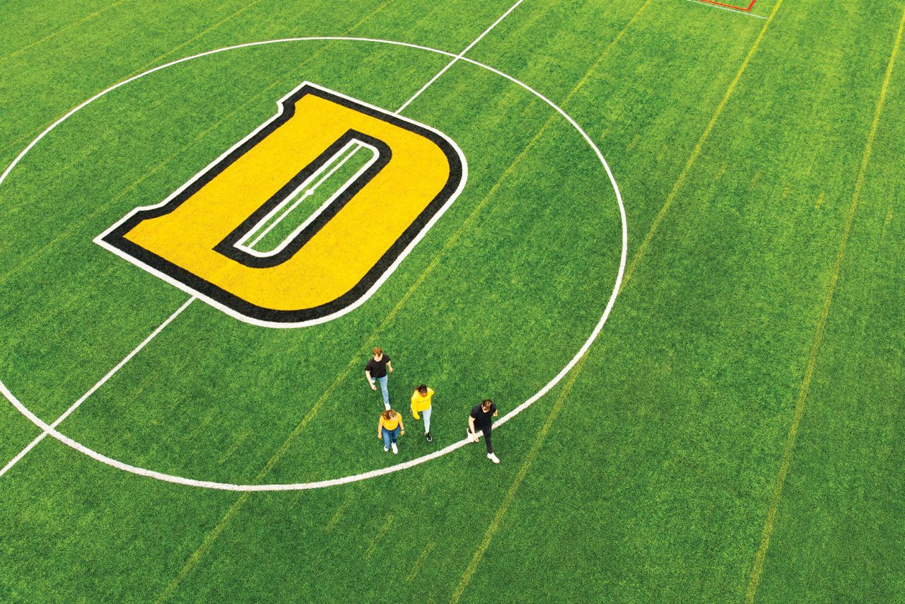 Aerial view of four students wearing black and yellow crossing green sports field painted with a large D surrounded by a white circle.