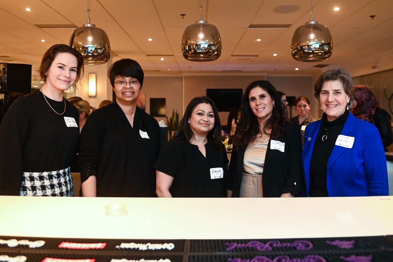 Five female-presenting women dressed in business casual attire wear nametags at an event in a restaurant setting.