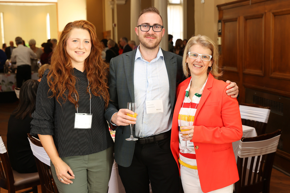 Three people hold Champagne glasses containing an orange drink at an event. They are dressed in business casual attire.