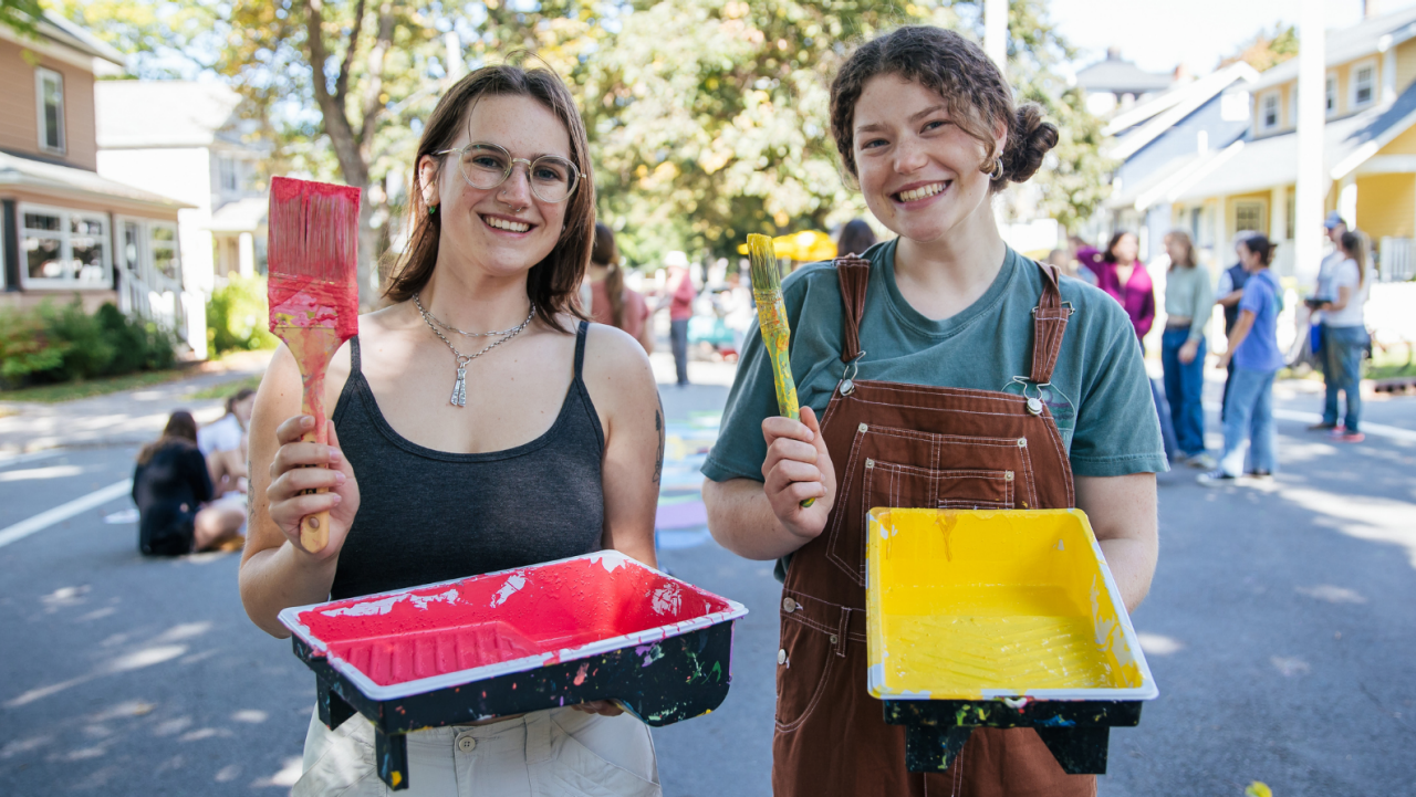Photo by Cody Turner, 2024 two female students holding paintbrushes and paint trays, laughing