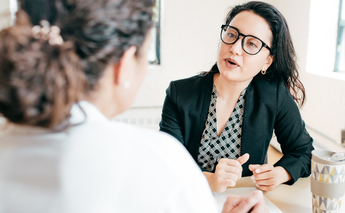 Two women sit face-to-face in conversation with a piece of paper and a take-out coffee on the table. 