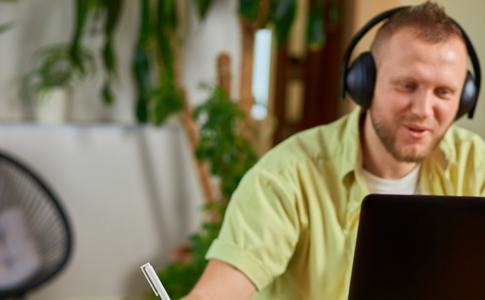 A man sits at a desk with a laptop and notebook. He wears headphones and is dressed casually.