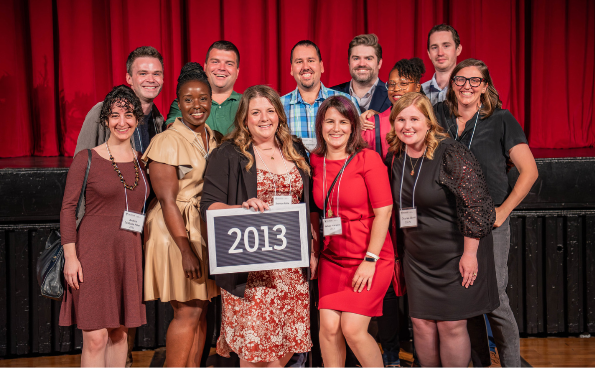 Twelve people stand in 2 rows dressed semi-formally in front of a stage with a red curtain. One holds a sign with “2013.”
