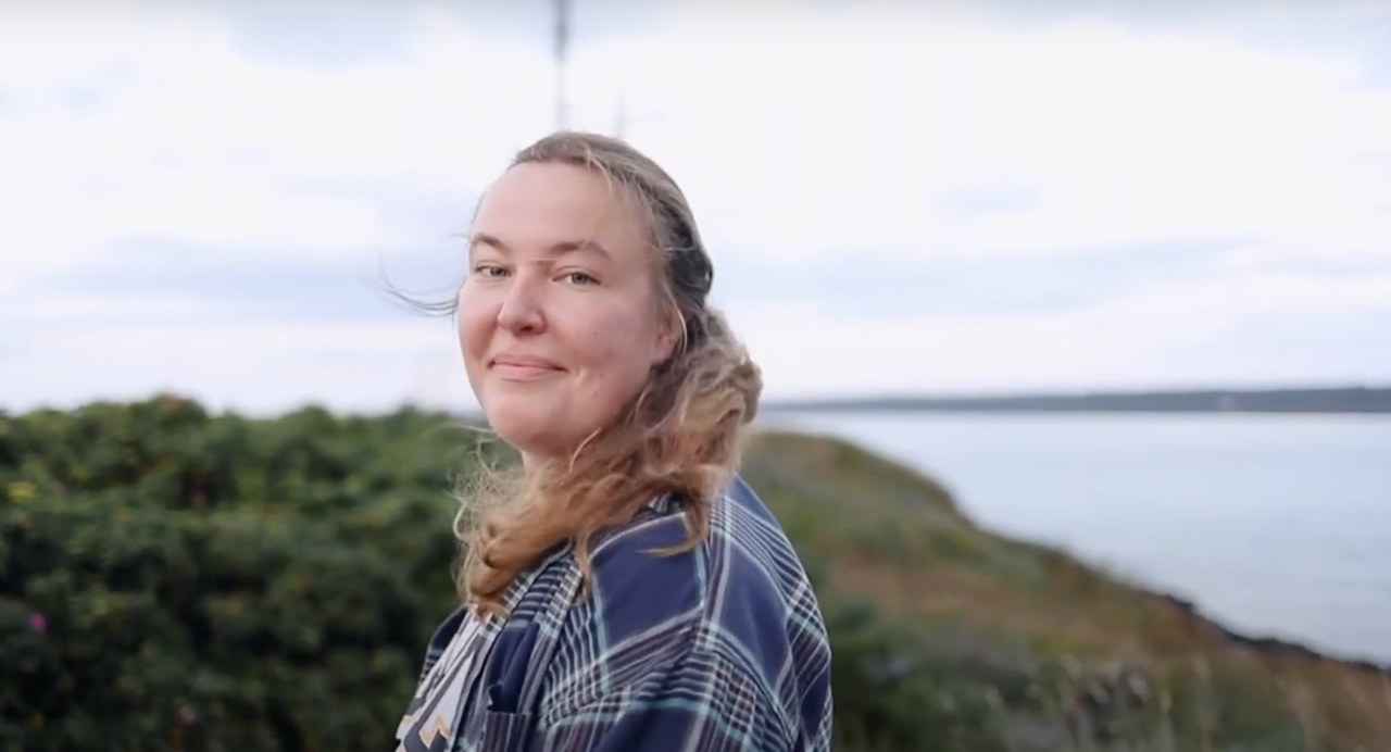 Female presenting student with long blond hair. She is standing outdoors with the ocean in the background.