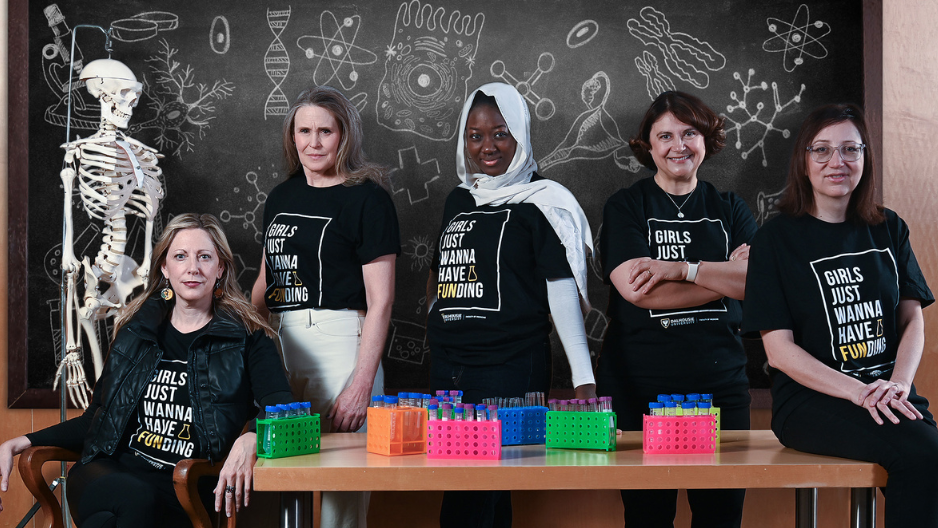 Five women in matching black t-shirts pose in a science classroom, surrounded by test tubes, a skeleton model, and a blackboard filled with science doodles. Three stand behind a desk, one sits in a chair, and another sits on the desk.
