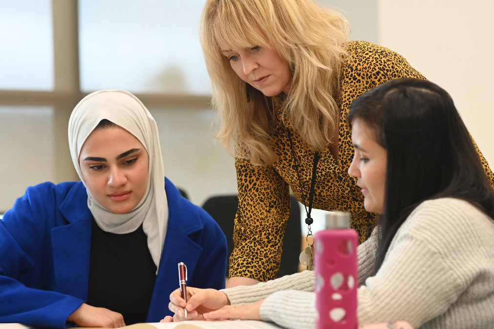 Two students at a desk work from a book while professor stands next to them.