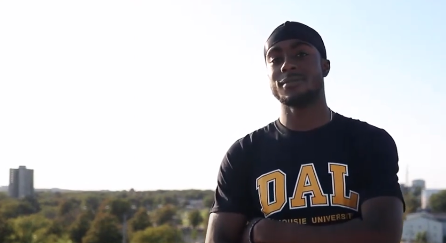 Male presenting Black student standing outside with arms crossed wearing Dal t-shirt.